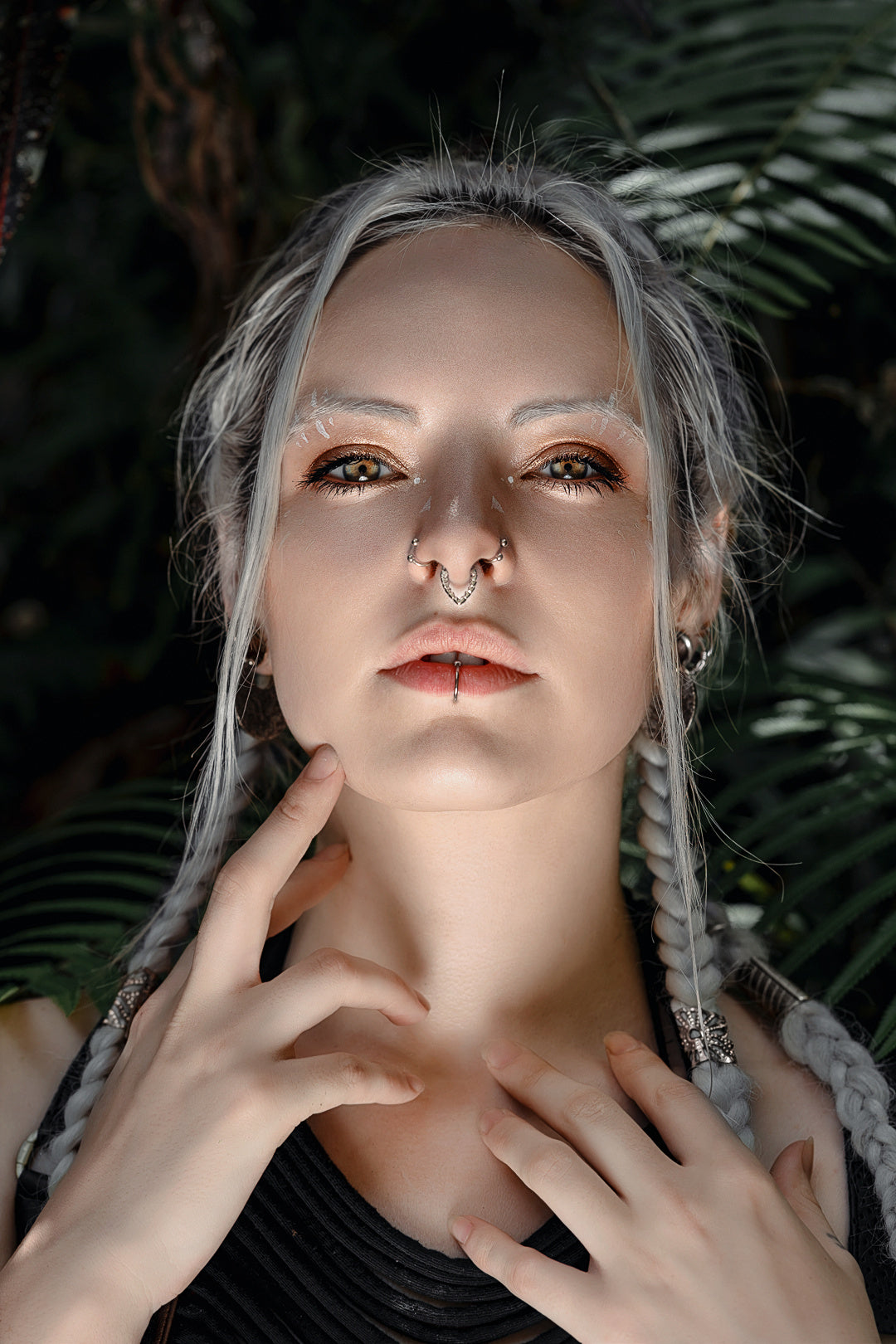 A person with light skin and blonde braids gazes intensely at the camera. They have facial piercings, including a striking Kaya Movement KAMO Biomech Teardrop Clicker Septum Ring in Silver, 16 gauge, and wear edgy makeup that highlights their cheekbones. Their hands lightly touch their chin and collarbone. Lush greenery forms the background.